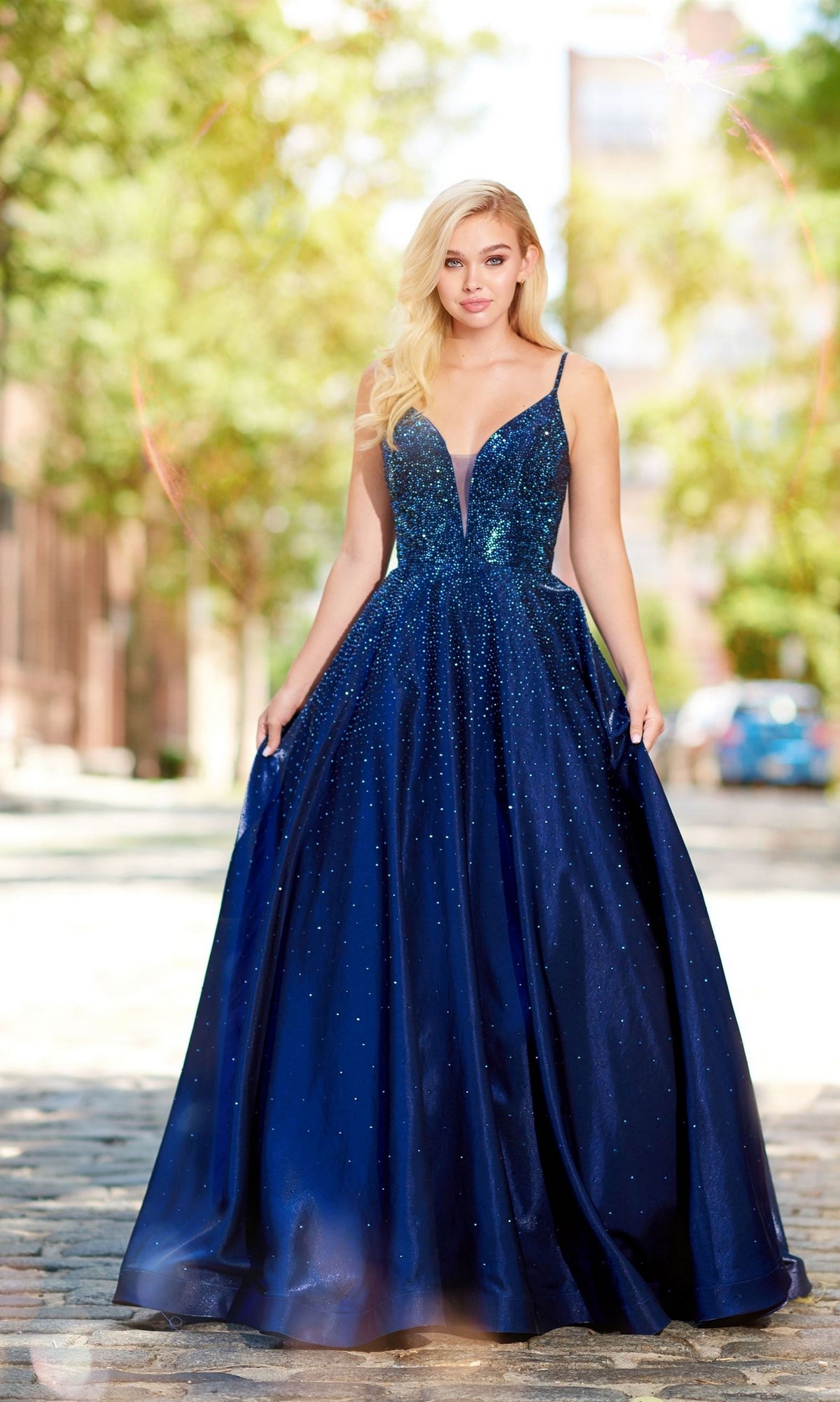 A woman stands outside on a cobblestone path, wearing the Ellie Wilde Ew121005 long beaded prom ball gown with a plunging neckline and sparkling embellishments.